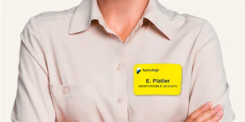 En person med lys beige skjorte med knapper og lett oppbrettede ermer. Personen har armene i kryss. På venstre side av brystet er en gul navnelapp med en blå og svart logo, teksten 'Technology', navnet 'E. Platier' og tittelen 'RESPONSABLE ACHATS' i svart skrift. Nøytral bakgrunn for tydelig synlighet.