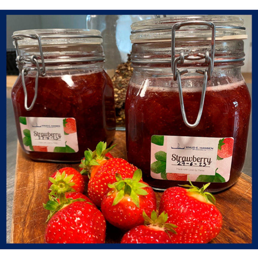 Syltetøyglass med jordbærmarmelade og en etikett med teksten Strawberry. Står på et trefat, omgitt av jordbær.