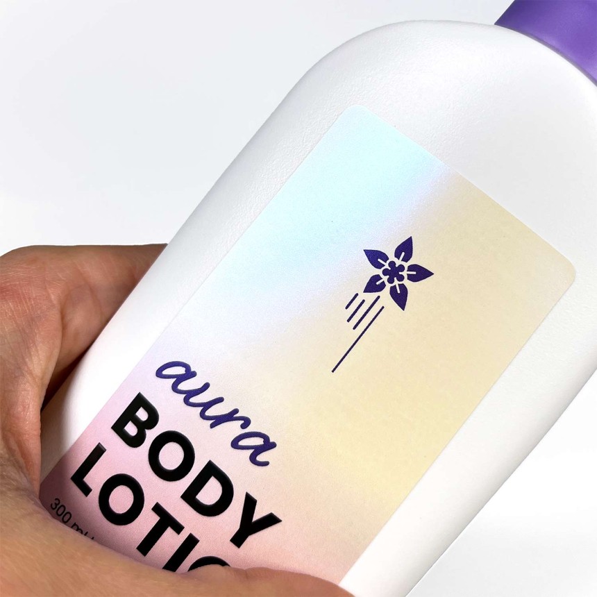 Etikett i perlemorsskinnende folie med teksten «aura body lotion». Her vises den glansfulle effekten på en hvit flaske.