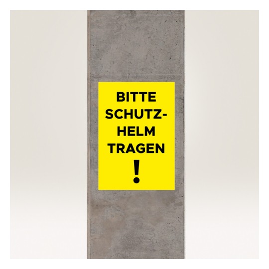 Bildet viser et gult skilt på en betongsøyle med teksten 'BITTE SCHUTZ-HELM TRAGEN!' som oversettes til 'Vennligst bruk hjelm!' og indikerer et sikkerhetskrav.