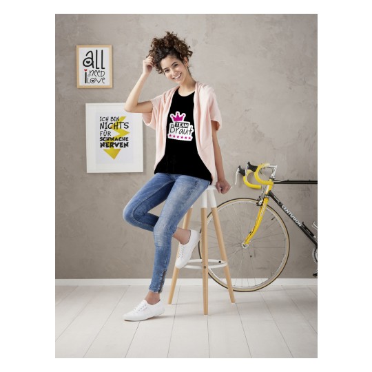 En ung kvinne sitter på en krakk, iført en svart t-skjorte med rosa detaljer som sier 'TEAM Braut'. Hun har krøllete hår og har på seg lyseblå jeans og hvite joggesko. En sykkel er synlig i bakgrunnen, sammen med noen innrammede kunstverk på veggen.