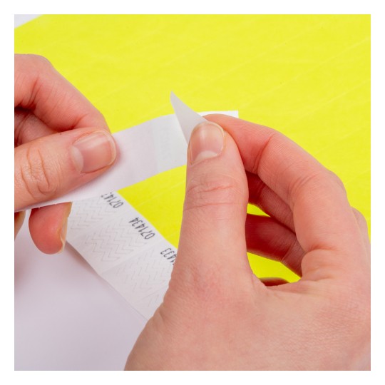 Bildet viser en persons hender som fjerner en papir- eller klistremerkestripe fra et ark, med en lys gul bakgrunn og trykte design på stripen.