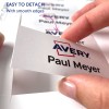 Bildet viser en hånd som løsner en etikett fra et ark, med navnet 'Paul Meyer' og 'Avery'-logoen, samt en merknad som indikerer at den er lett å løsne med glatte kanter.
