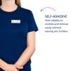 Bildet viser en person med en marineblå skjorte med et navneskilt som lyder 'B. Carlos, markedsdirektør.' Det er også en beskrivelse som sier at navneskiltet er selvklebende og kan festes på klær pålitelig uten å etterlate rester.