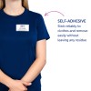 Bildet viser en person som har på seg en marineblå t-skjorte med et selvklebende navneskilt som lyder 'B. Carlos, Markedsføringsdirektør.' Ved siden av dem er det en beskrivelse som fremhever den selvklebende egenskapen til navneskiltet, med vekt på dets pålitelighet og letthet ved fjerning uten rester.