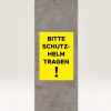 Bildet viser et gult skilt på en betongsøyle med teksten 'BITTE SCHUTZ-HELM TRAGEN!' som oversettes til 'Vennligst bruk hjelm!' og indikerer et sikkerhetskrav.