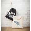 Bildet viser to vesker mot en hvit murvegg. En svart snørepose med ordene 'Love, Peace, Party' henger der. Ved siden av vises en beige handlenettveske med et dekorativt fjærdesign.